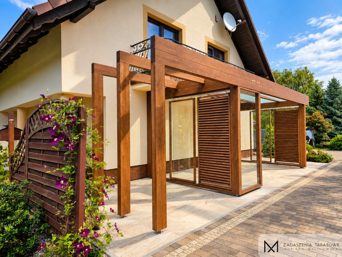 Pergola tarasowa z drewna BSH połączona ze szklanym zadaszeniem i przeszkleniami bocznymi w kolorze orzech.